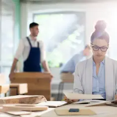 Kobieta w okularach analizuje dokumenty przy biurku. W tle mężczyzna w fartuchu. To może być staż z urzędu pracy dla osób długotrwale bezrobotnych, realizowany w ramach specjalnych programów celowych.