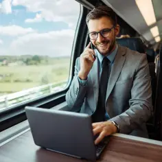 Uśmiechnięty biznesmen w garniturze rozmawia przez telefon i pracuje na laptopie w pociągu. Czy wyjazd na szkolenie to delegacja i czy pracownikowi należy się dieta?