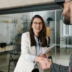 Uśmiechnięta kobieta z dokumentami w ręku ściska dłoń mężczyzny. Jak wytłumaczyć częste zmiany pracy rekruterowi by nie wyjść na skoczka?
