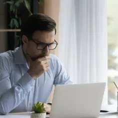 Mężczyzna w okularach siedzi przy biurku i myśli, patrząc na laptopa.