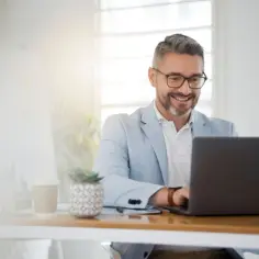 Uśmiechnięty mężczyzna w jasnej marynarce pracuje przy laptopie. Jego podsumowanie zawodowe na samej górze CV to klucz do sukcesu, jak w 3 zdaniach uwieść HR.
